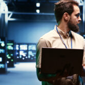 Supervisor using laptop to oversee energy consumption across server hub components and distributors. Serviceman ensuring high tech facility sensors for temperature and humidity are working properly